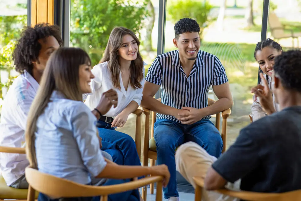 A group of stylish adults sit in wooden chairs in a circle while participating in group therapy