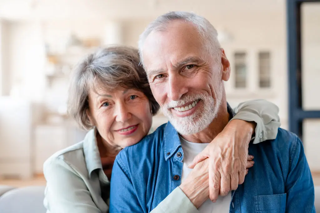 An older woman embraces her husband from behind as they both smile